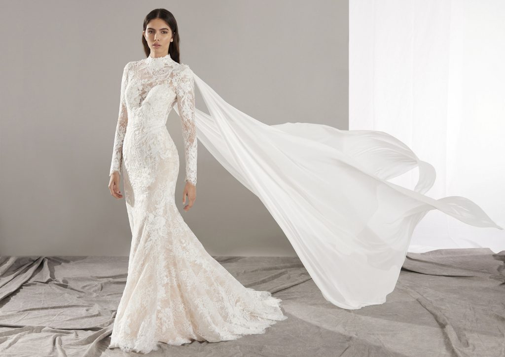 Bruid in zeemeermin-trouwjurk van kant met hooggesloten hals en zwierige cape.