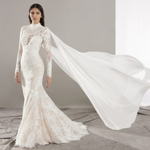 Bruid in zeemeermin-trouwjurk van kant met hooggesloten hals en zwierige cape.