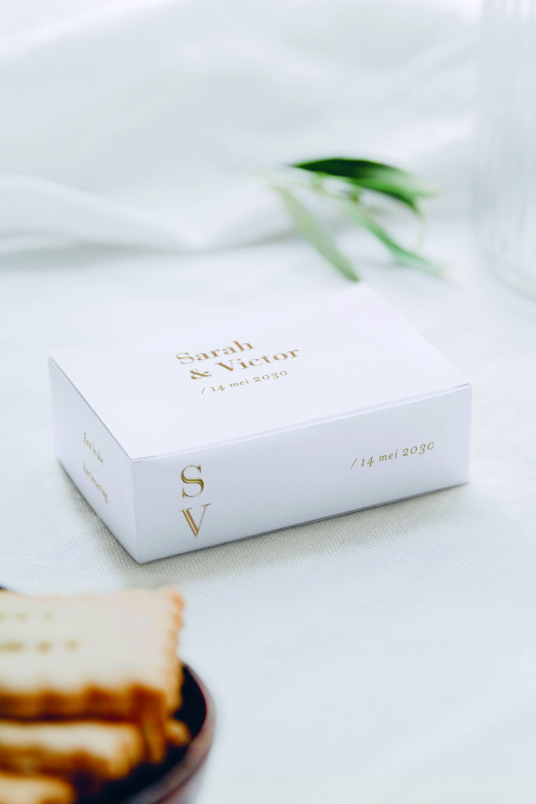 Elegante witte save-the-date-verpakking met gouden letters, met de namen "Sarah & Victor" en de trouwdatum "14 mei 2030", geplaatst op een lichte, sfeervolle tafelsetting met koekjes en olijfgroen als decoratie