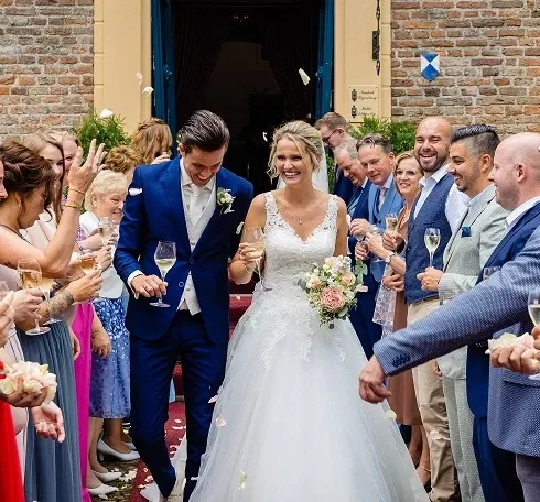 Bruid en bruidegom vieren feestelijk toostmoment met gasten buiten bij kasteelmuur, omringd door vrienden en familie bij kasteel Wijenburg.
