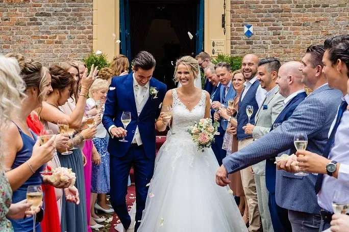 Bruid en bruidegom vieren feestelijk toostmoment met gasten buiten bij kasteelmuur, omringd door vrienden en familie bij kasteel Wijenburg.