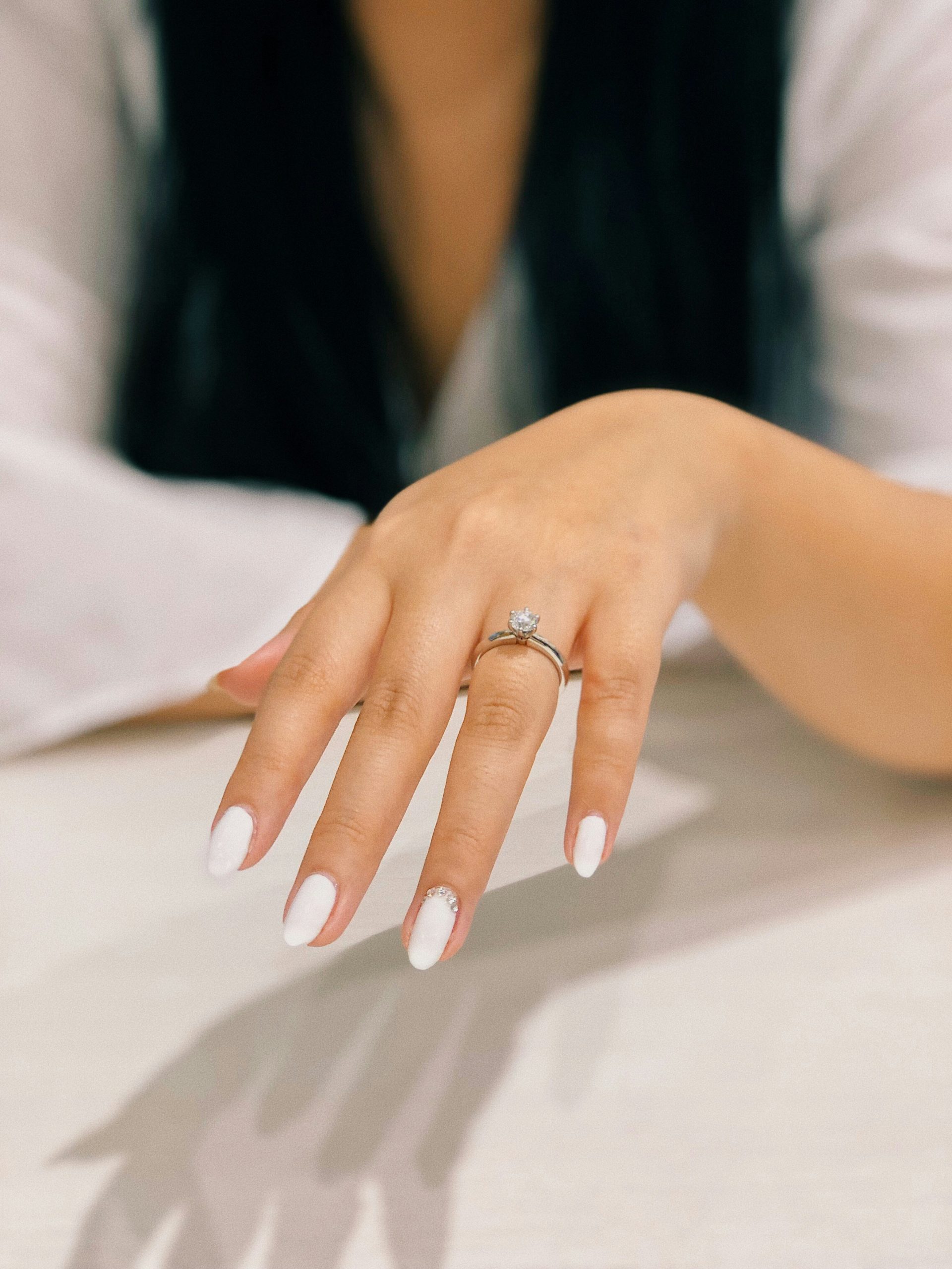 Een close-up van een vrouwelijke hand met een elegante witte manicure en een zilveren verlovingsring met diamant, rustend op een lichte tafel.