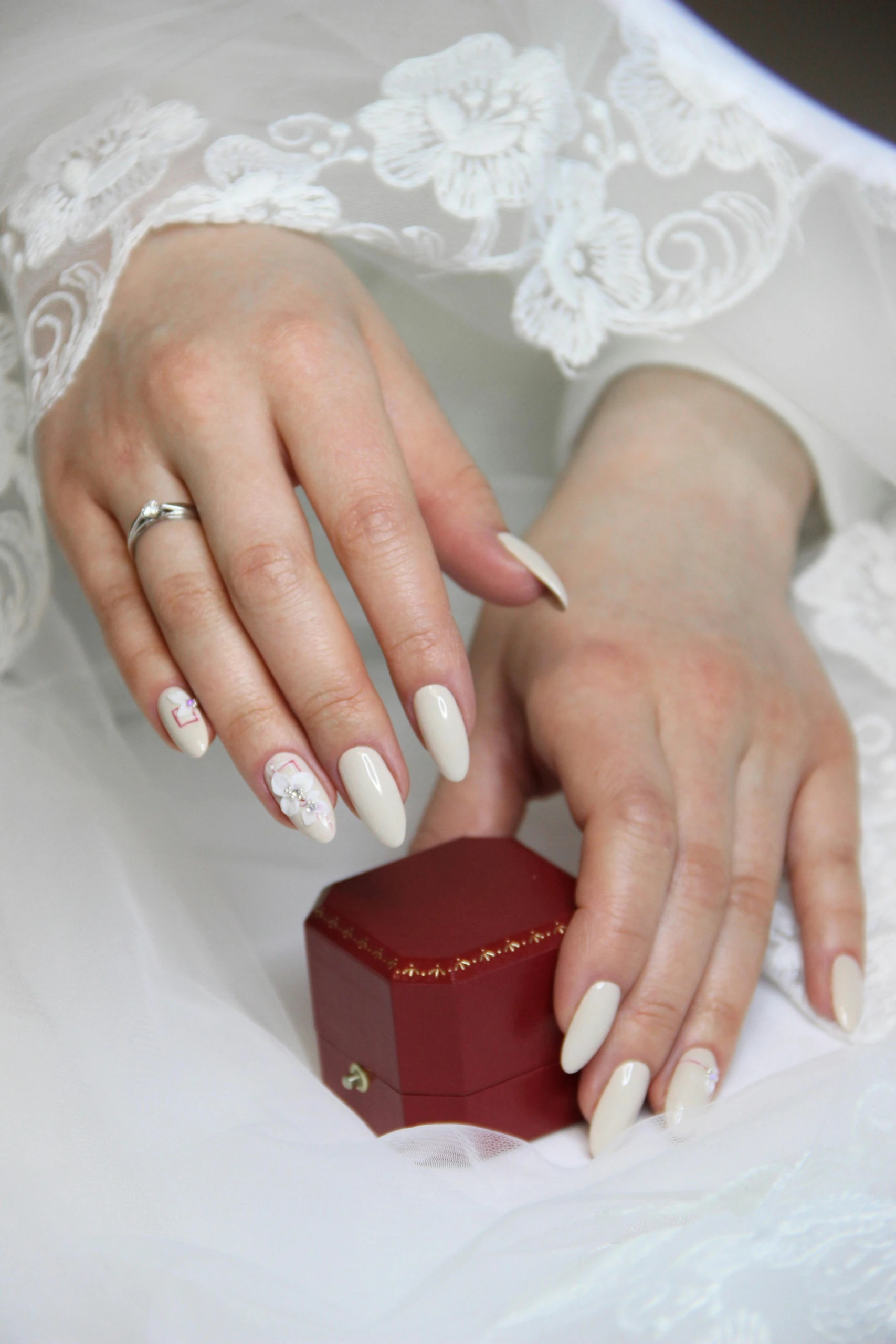 Een close-up van een bruid met elegante, amandelvormige nude-gel nagels met bloemendecoratie en een zilveren verlovingsring, rustend op een rood sieradendoosje.