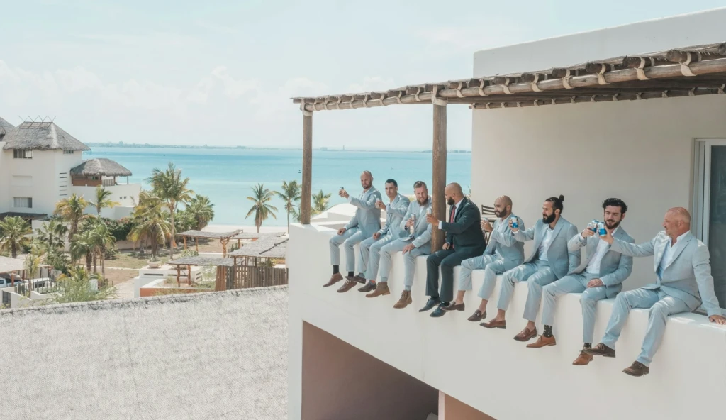Een groep mannen in lichtblauwe pakken en bruidegom in een donker trouwpak, zitten op de rand van een balkon met uitzicht op een prachtig strand en de oceaan, terwijl ze lachen en blikjes drinken.