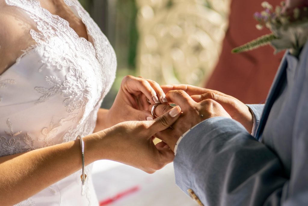 Close-up van bruid die trouwring om de vinger van haar partner schuift tijdens de ceremonie, met elegante kanten trouwjurk en zonnige achtergrond.