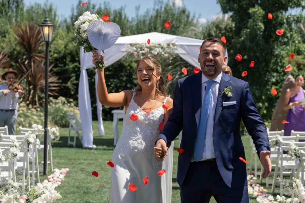 Bruidspaar loopt hand in hand lachend onder vallende rozenblaadjes na hun ceremonie in de buitenlucht, met witte bloemen en een boog op de achtergrond.