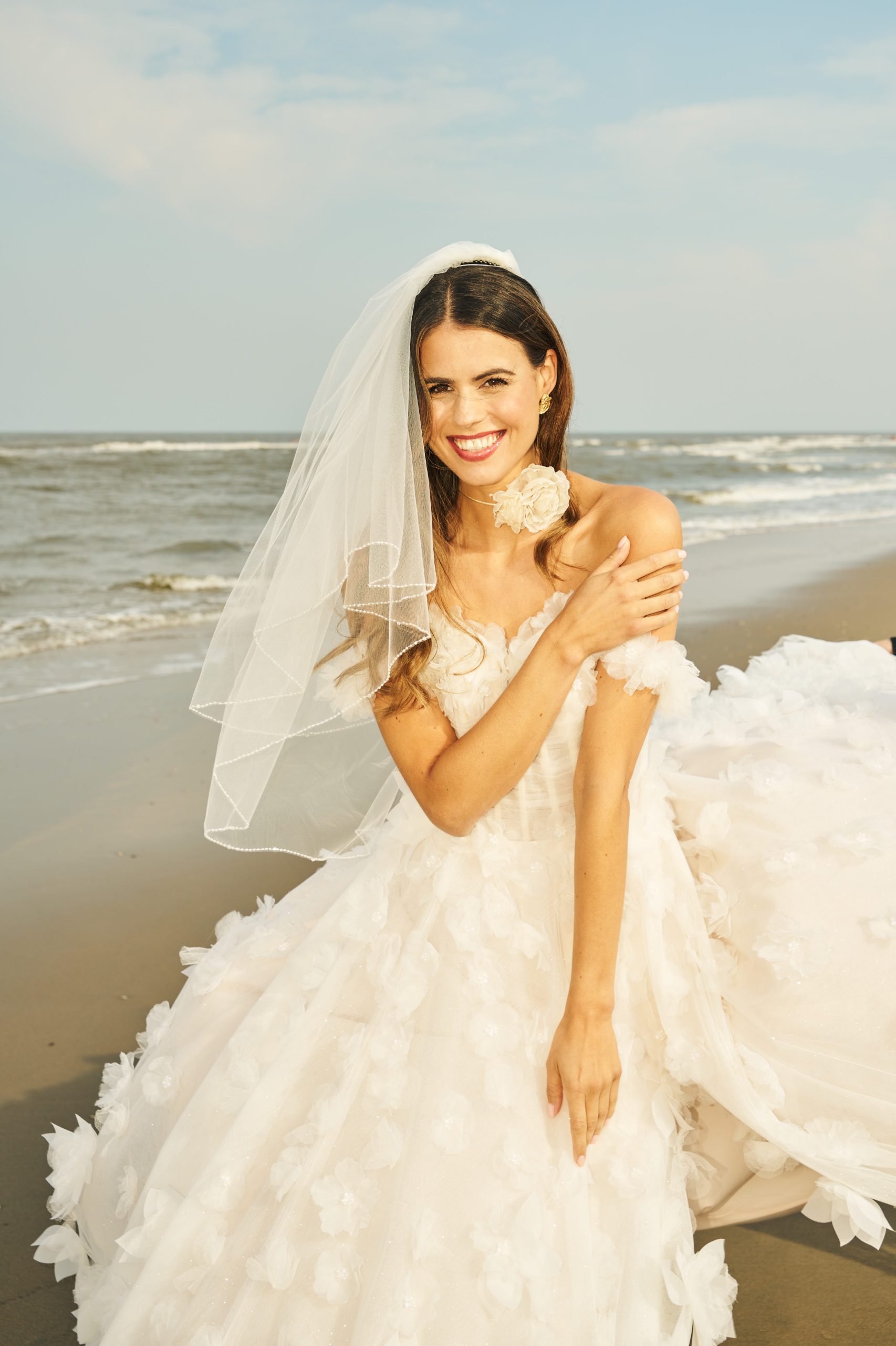 Een stralende bruid poseert op het strand in een romantische prinsessenjurk met driedimensionale bloemenapplicaties en een luchtige sluier. De zachte zeewind en het gouden zonlicht versterken de speelse, dromerige sfeer van deze zomerse bruiloft.