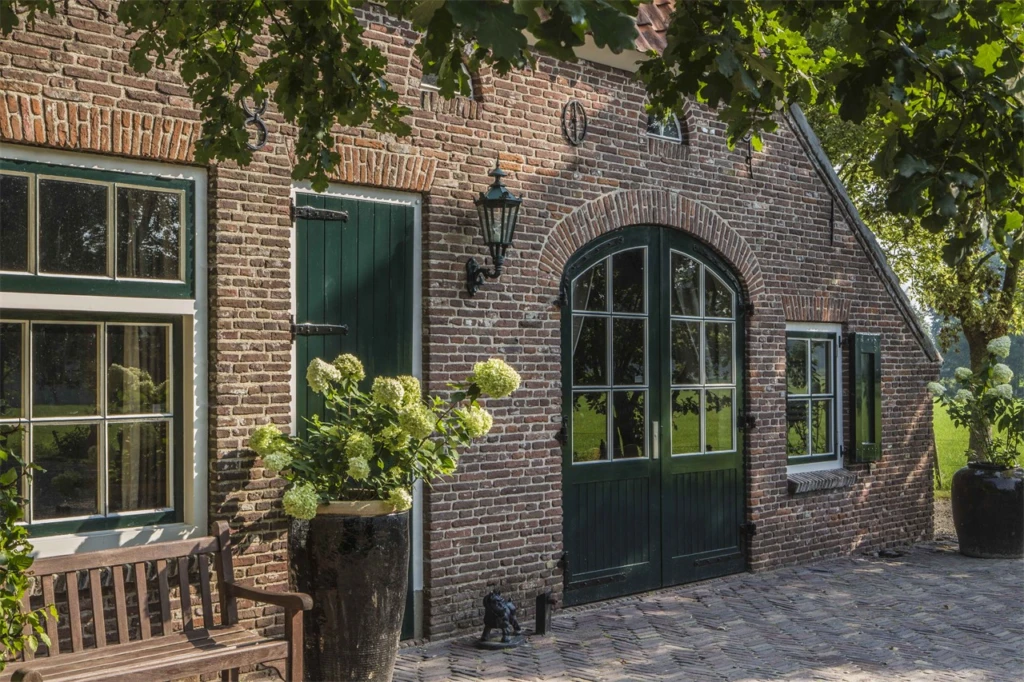 Een rustieke boerderij met bakstenen muren, groene houten deuren en ramen, omringd door een natuurlijke omgeving met hortensia's in grote potten en een houten bank op een geplaveide ondergrond. Een sfeervolle landelijke setting