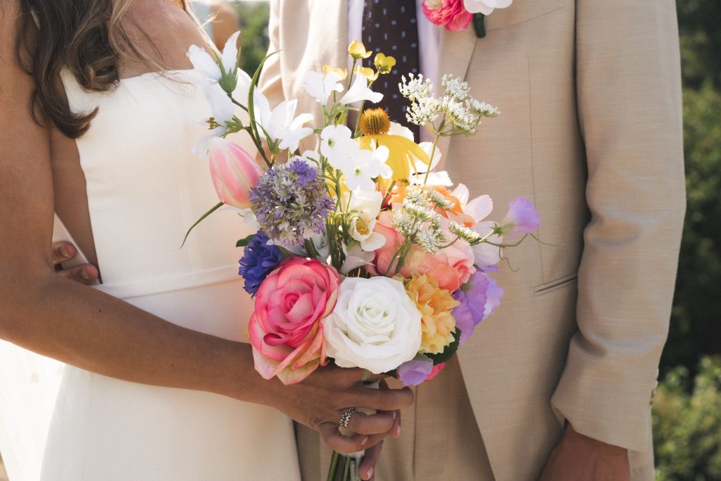 Een bruid houdt een kleurrijk zijden bruidsboeket vast met roze rozen, witte en paarse bloemen, en zachte groene accenten. De bruidegom, gekleed in een beige pak met bijpassende corsage, staat naast haar. Het boeket straalt elegantie en tijdloze schoonheid uit.