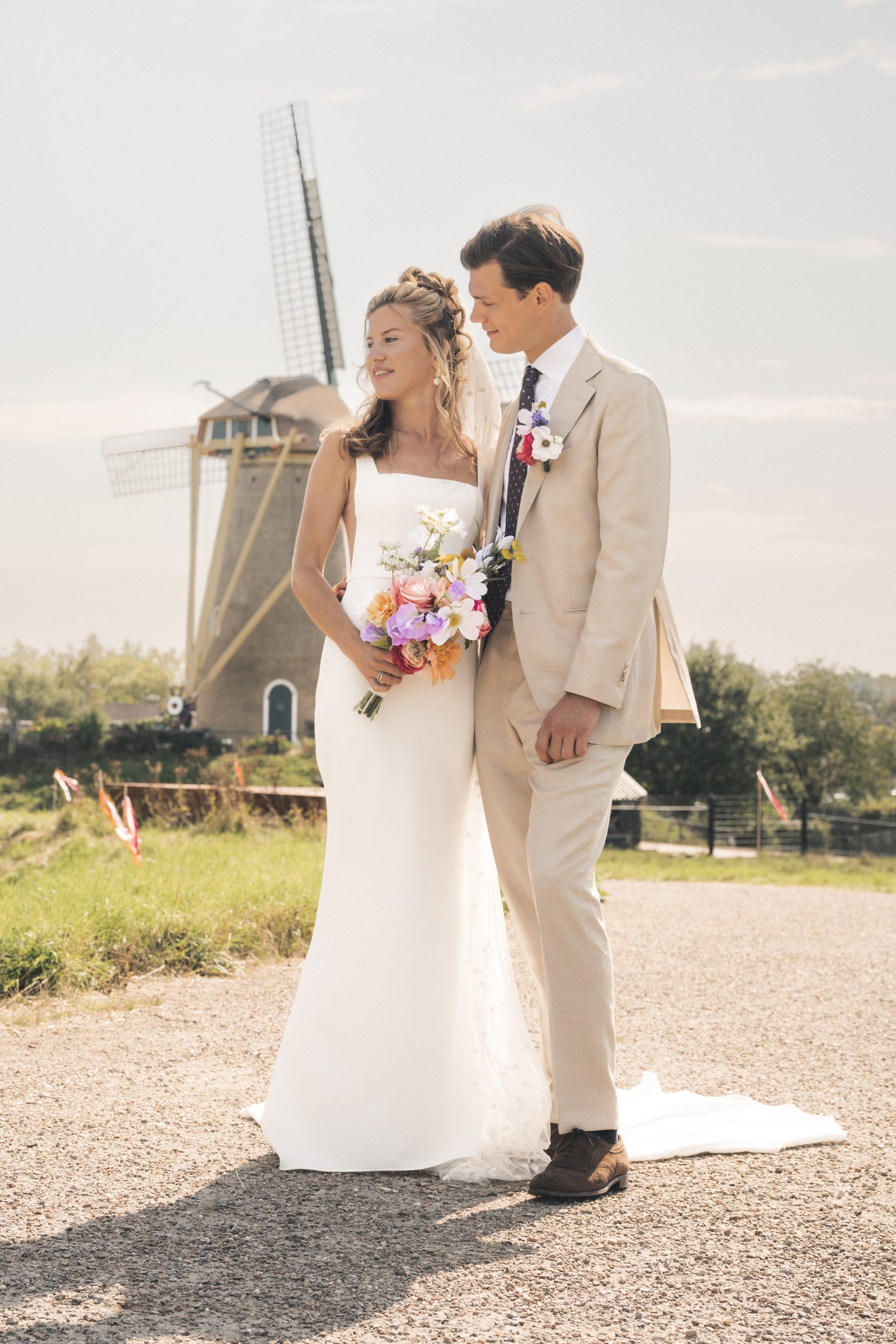 Een bruid en bruidegom staan samen voor een traditionele windmolen. De bruid draagt een elegante witte jurk en houdt een kleurrijk zijden boeket vast, terwijl de bruidegom in een beige pak met bijpassende corsage naast haar staat. Een idyllisch plaatje vol liefde en traditie.