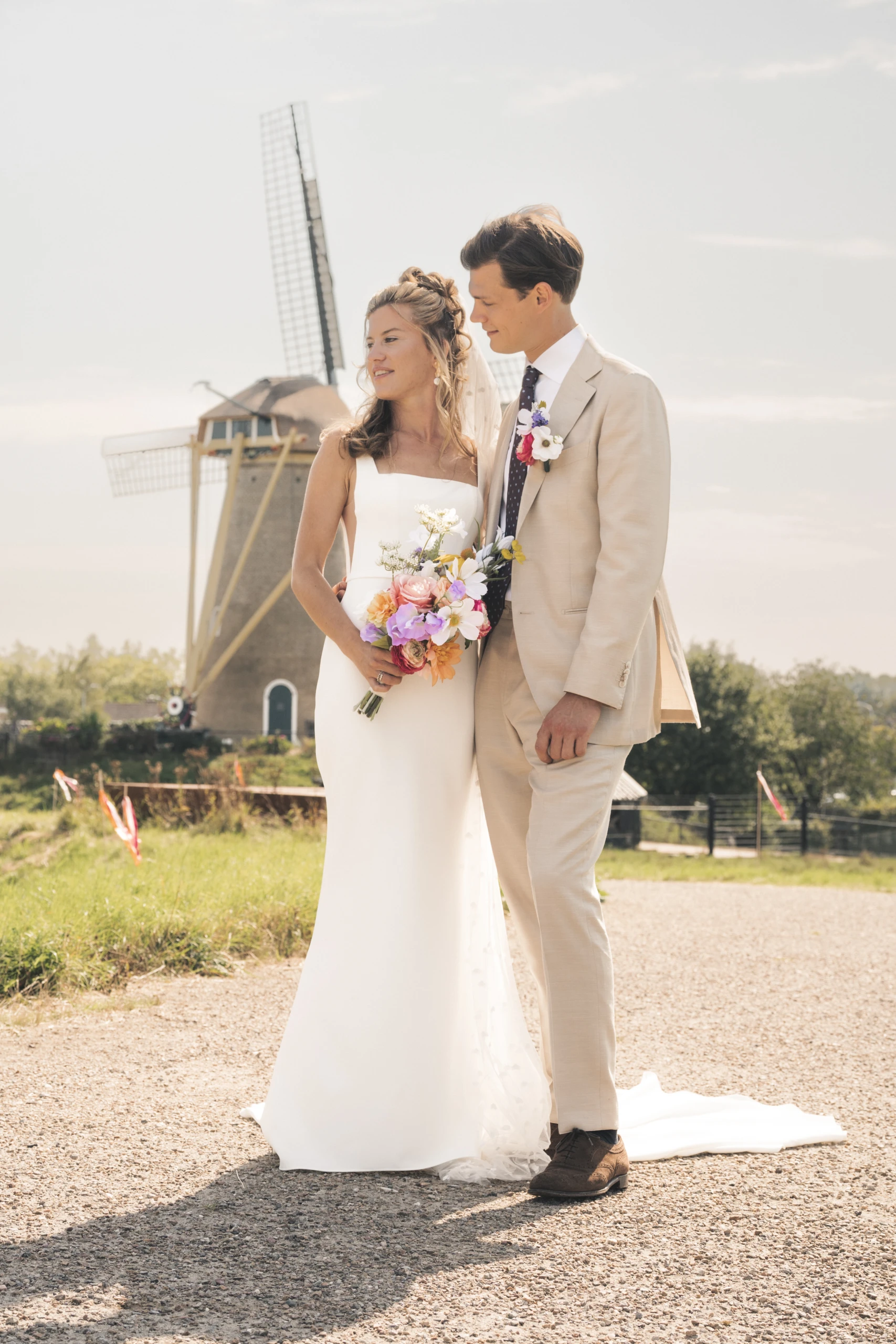 Een bruid en bruidegom staan samen voor een traditionele windmolen. De bruid draagt een elegante witte jurk en houdt een kleurrijk zijden boeket vast, terwijl de bruidegom in een beige pak met bijpassende corsage naast haar staat. Een idyllisch plaatje vol liefde en traditie.