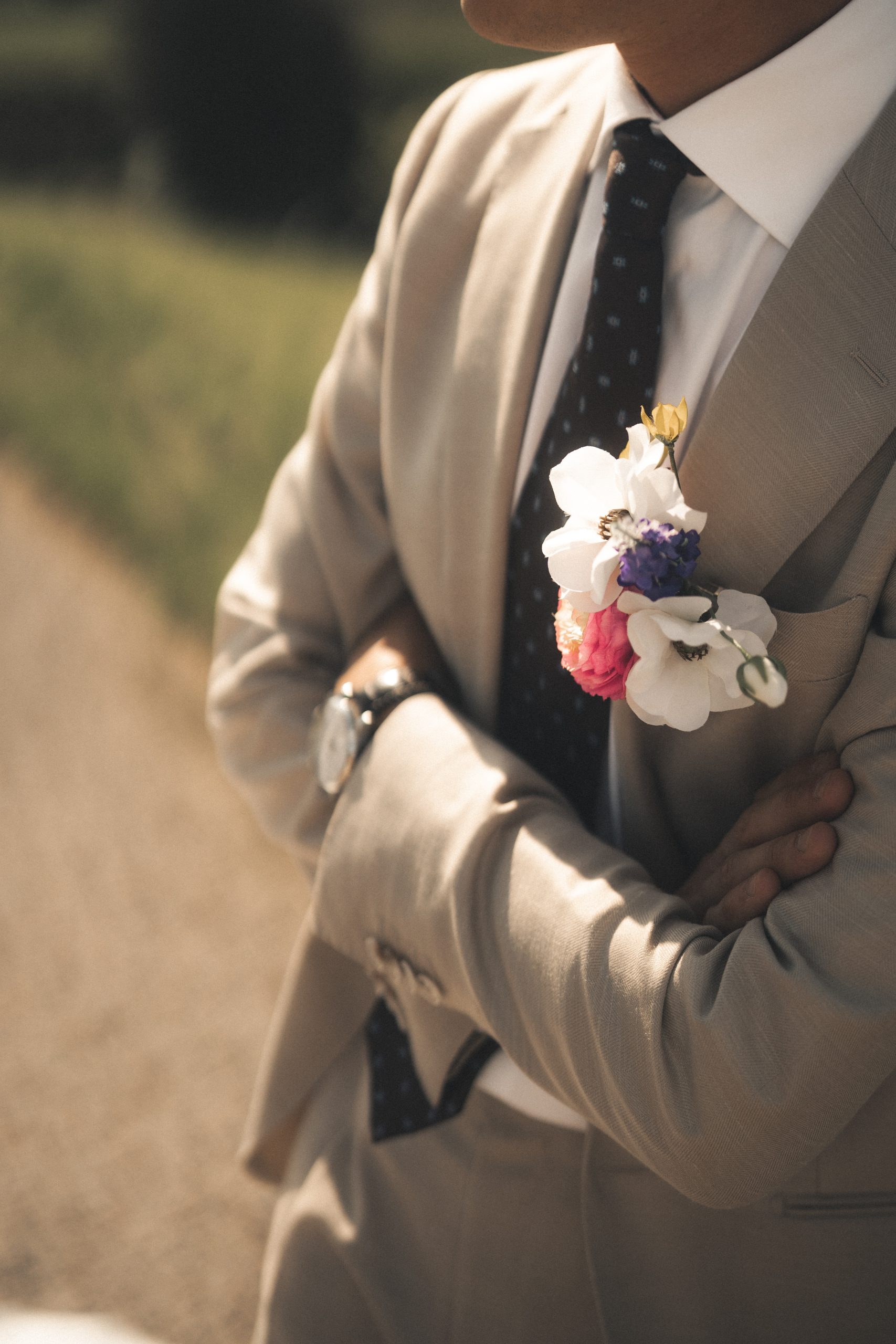 Close-up van de bruidegom in een beige pak met een stijlvolle zijden corsage op zijn revers. De corsage bevat witte, paarse en roze bloemen en vormt een perfecte match met het bruidsboeket. Zijn gekruiste armen en nette uitstraling geven een elegante en zelfverzekerde look.