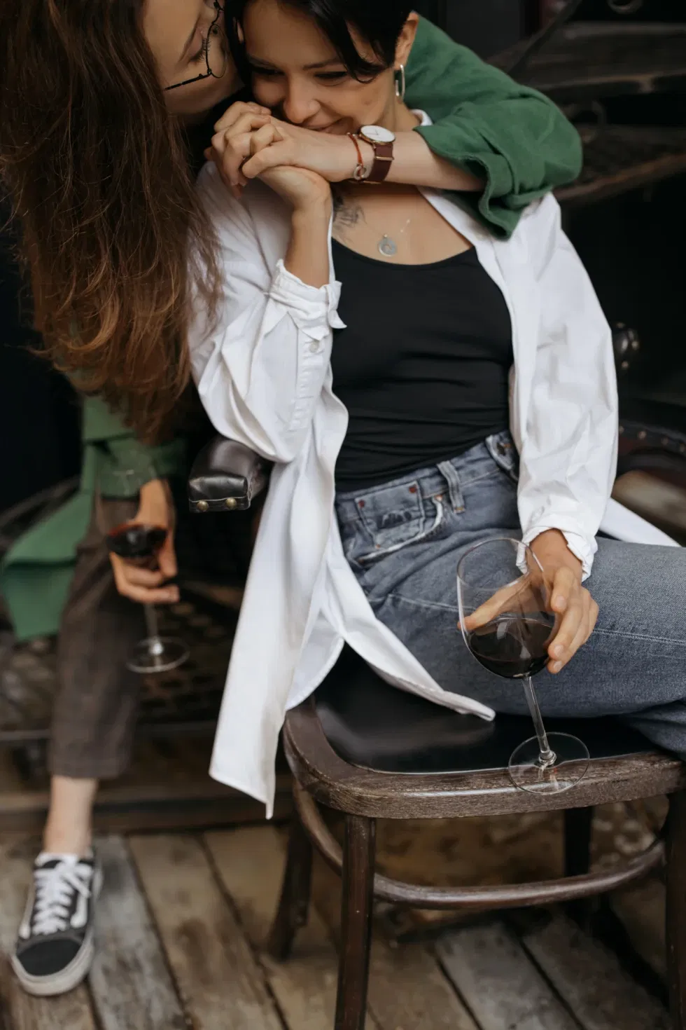 vrouw kust van achteren een andere vrouw. Ze houden elkaars hand vast. in de andere hand houden ze een wijnglas vast