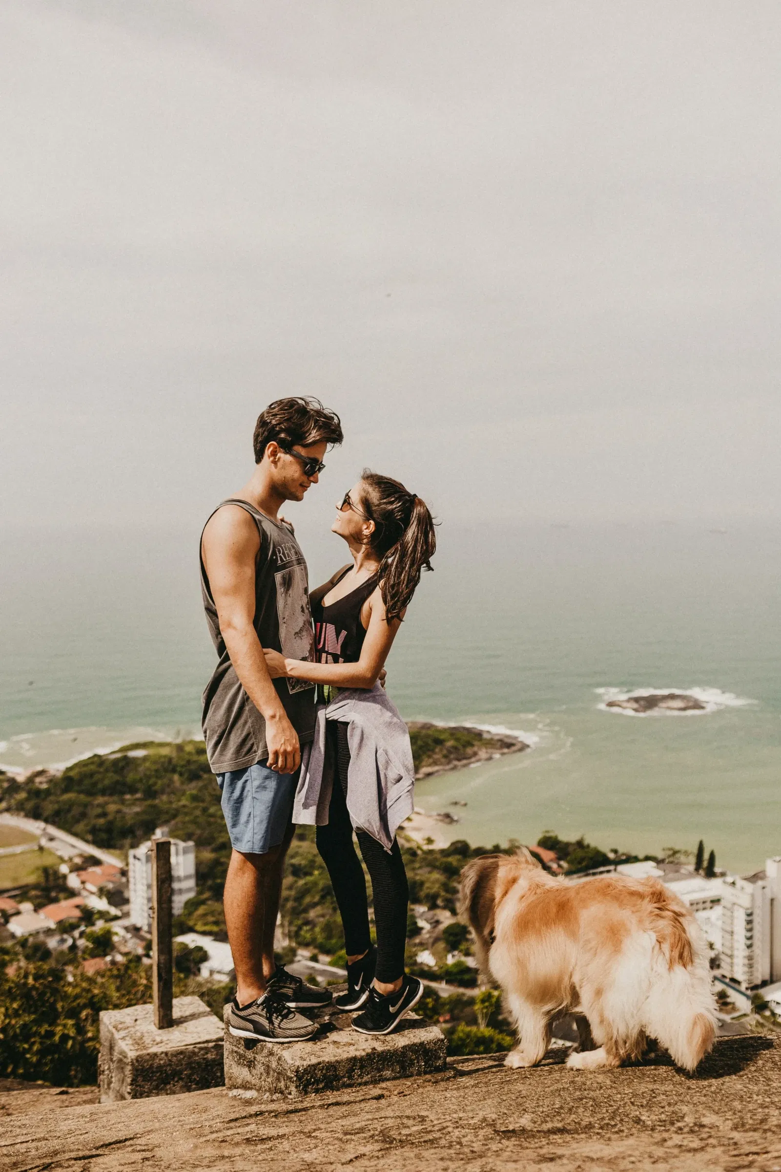 Stel in sportieve kleding kijken elkaar aan op de top van een berg met op de achtergrond de zee. Op de voorgrond staat een hond
