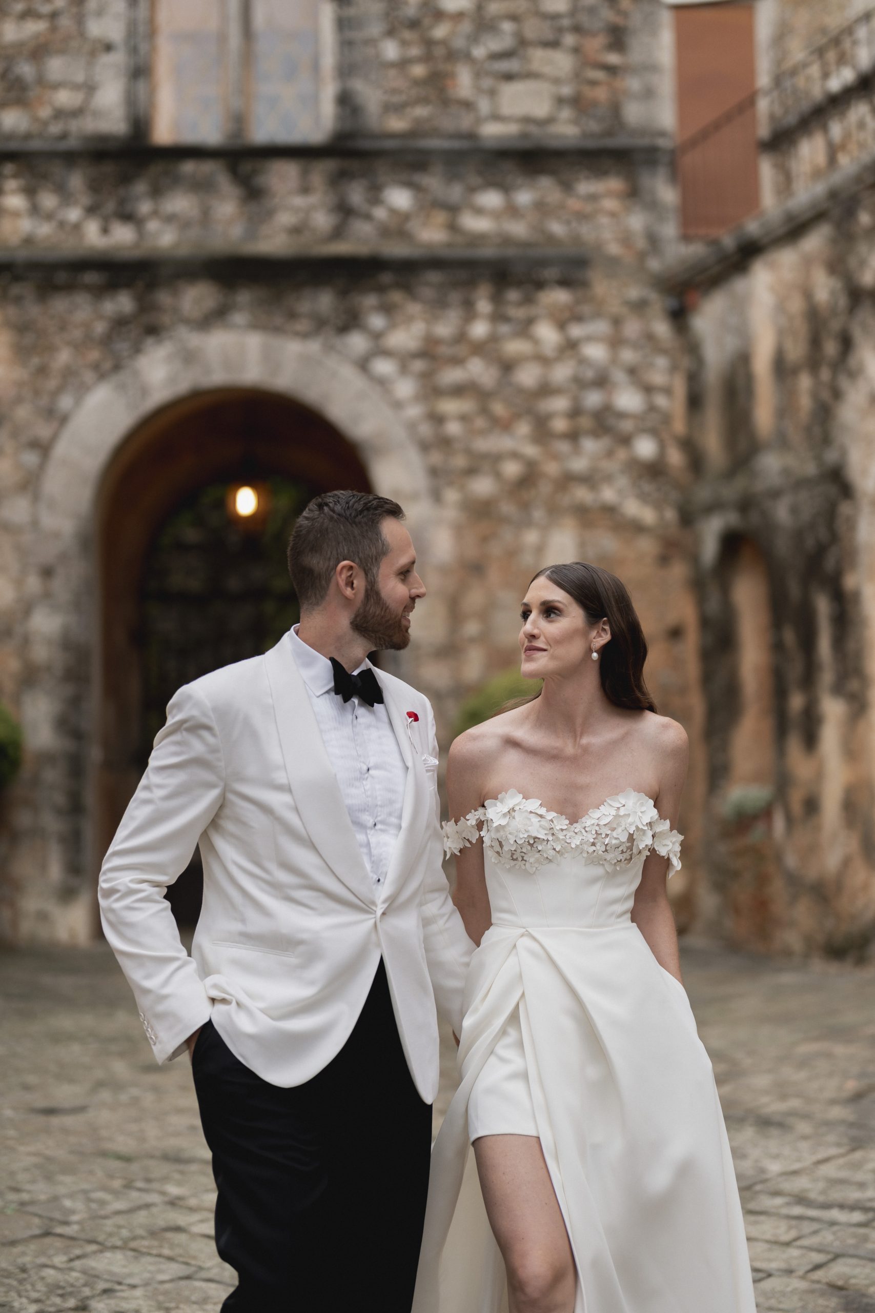 Bruidsmode CEO Justin Alexander en vrouw Kelsey Turchy in trouwkleding. Wit pak en witte jurk met hoge split en off the shoulder mouwtjes.