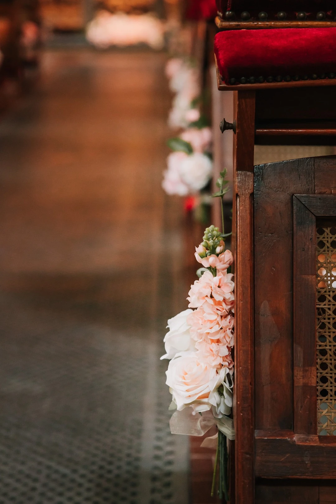 Close-up van een kerkbank versierd met een elegante bloemendecoratie van zijden rozen en pastelkleurige bloemen. De houten bank met rood fluwelen kussen voegt een klassieke en sfeervolle touch toe aan de bruiloftssetting. Op de achtergrond is het gangpad subtiel verlicht met meer bloemendecoraties.