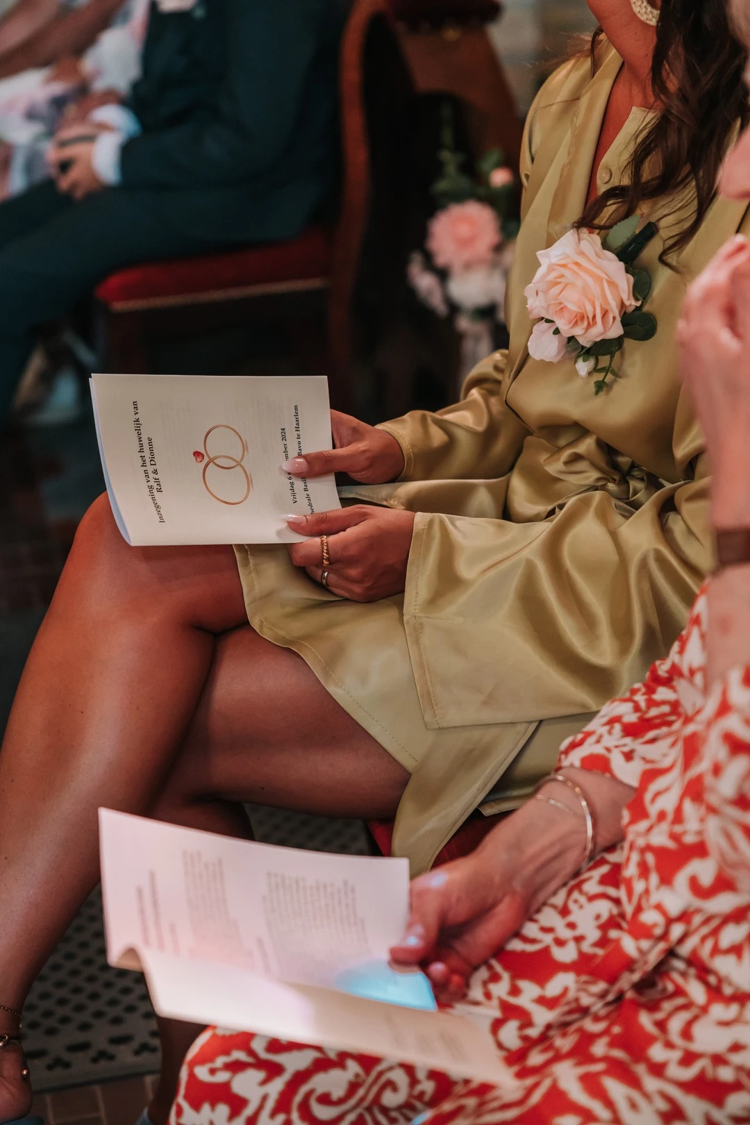 Een elegante vrouwelijke gast, gekleed in een satijnen jurk met een zachte groene tint, houdt een ceremonieboekje vast tijdens een bruiloftsdienst. Ze draagt een delicate zijden corsage met een perzikroze roos. Op de achtergrond zijn andere gasten en bloemversieringen op de kerkbanken zichtbaar, wat bijdraagt aan de romantische sfeer van de ceremonie.