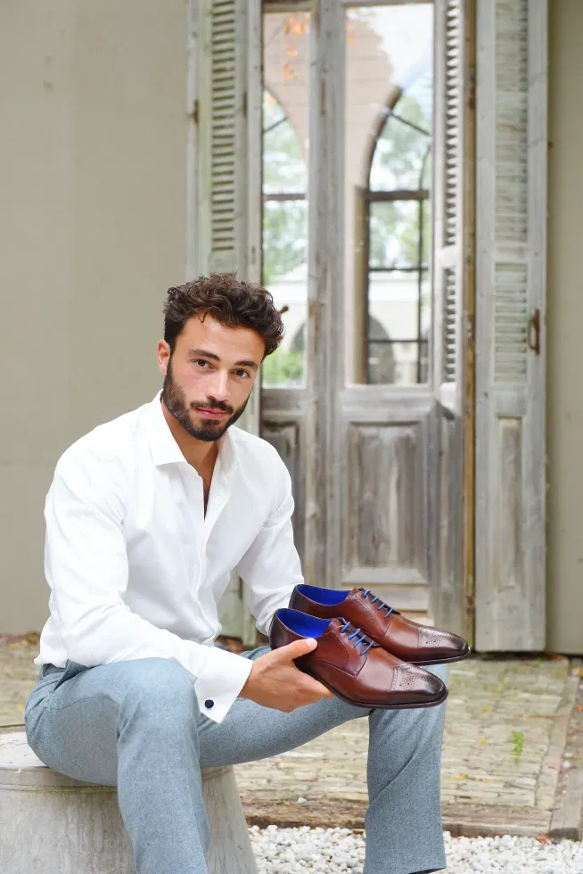 Man zit op een waterton in wit overhemd en lichtblauwe pantalon met twee leren schoenen met blauwe binnenkant en blauwe veters