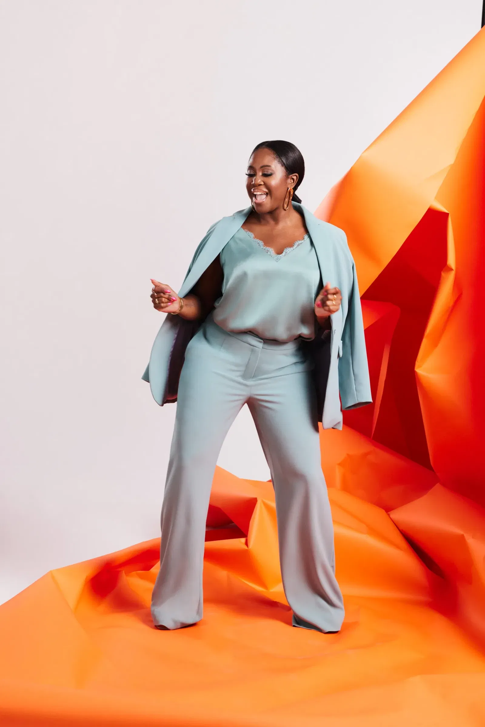 Vrouw staat op oranje papier in een blauw-grijs cocktailpak met een jasje over haar schouders