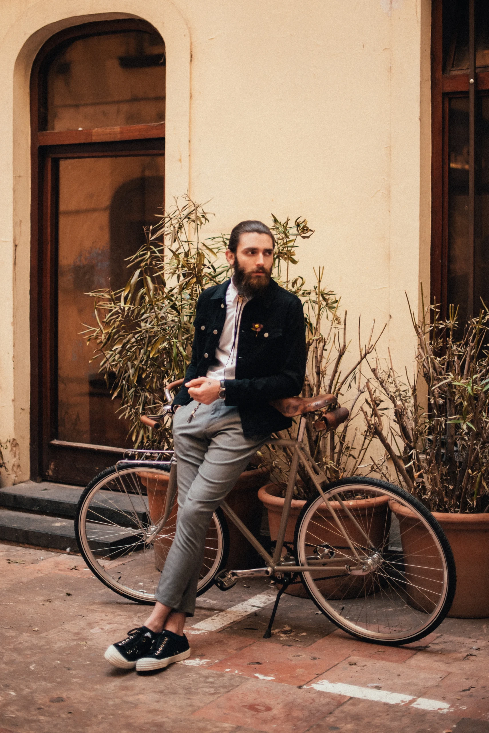 Man met baard en achterover gekamde haren leunt op een retro fiets met bruine zadel en kleine fietsendrager voorop.