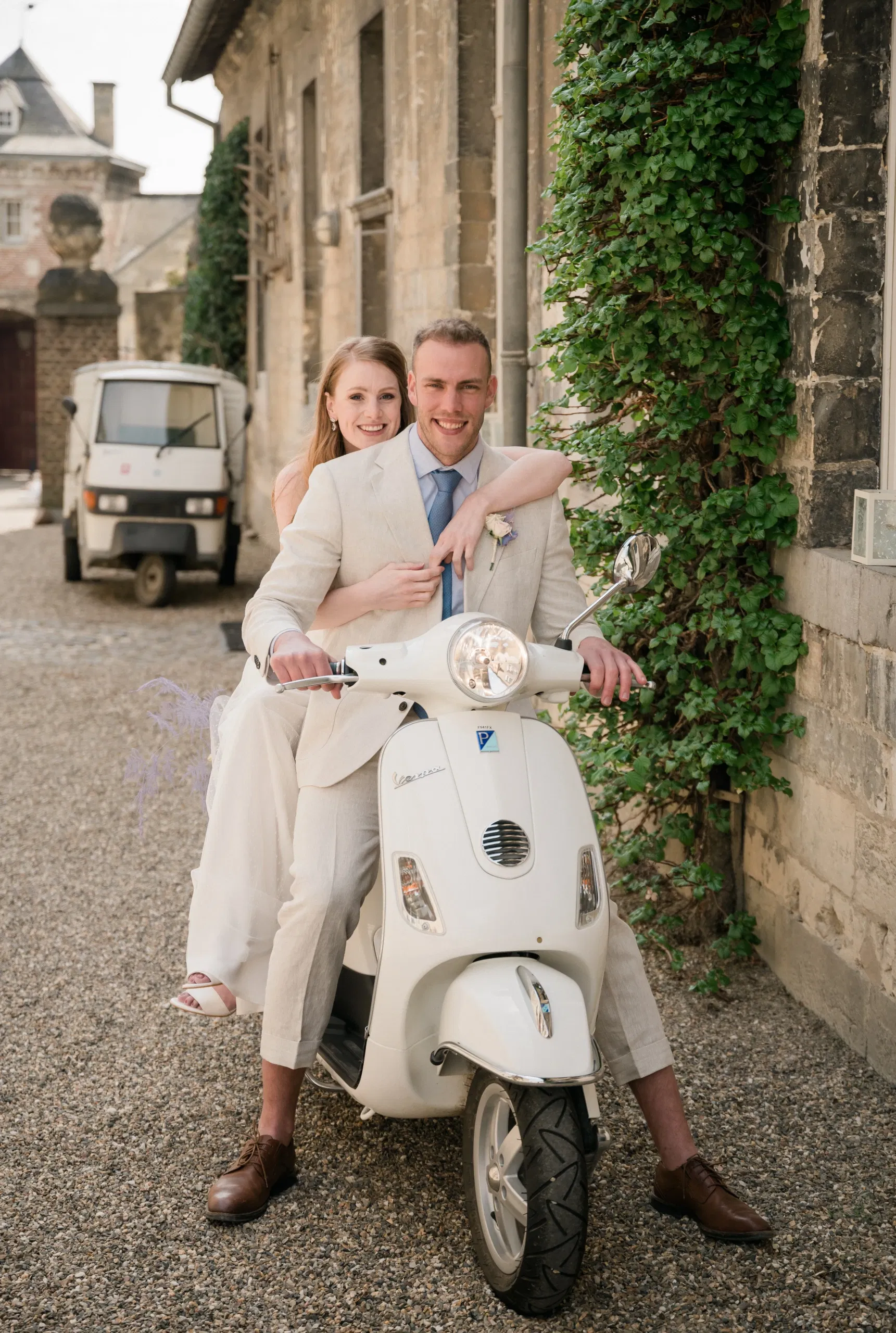Bruid zit achterop op een witte scooter bij de bruidegom. Bruidegom heeft een licht zandkleurig trouwpak aan met bruine schoenen. Bruid heeft een witte trouwjurk en trouwschoenen aan. Op de achtergrond een driewieler Piaggio