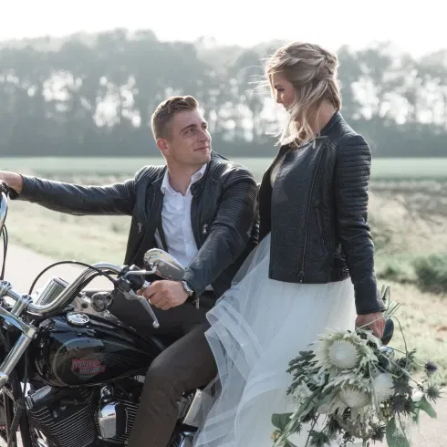 Bruidspaar kijkt elkaar aan zittend op een zwarte Harley Davidson. Beiden dragen een zwarte leren jas. Bruid in witte bruidsjurk houdt een wit bruidsboeket vast