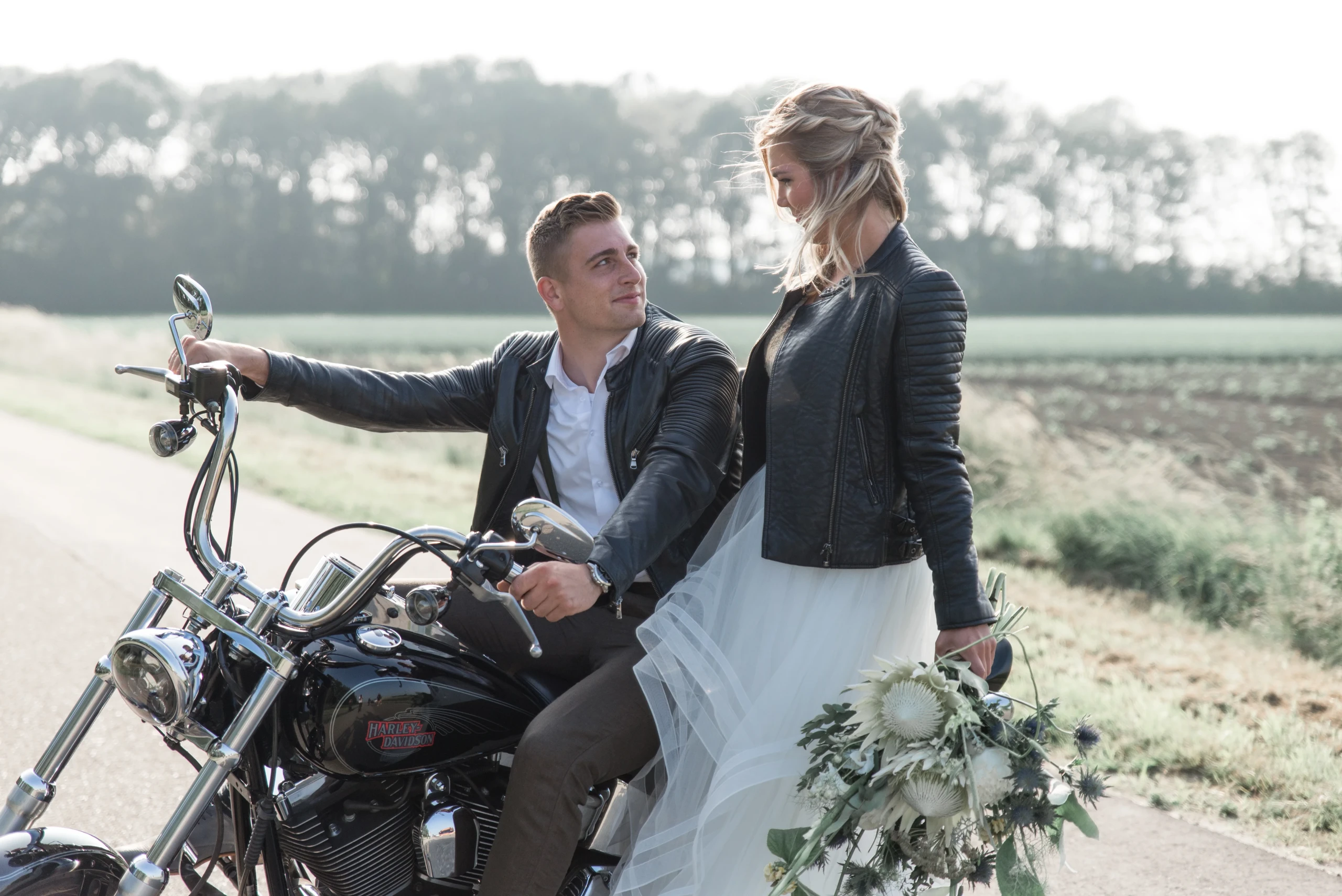 Bruidspaar kijkt elkaar aan zittend op een zwarte Harley Davidson. Beiden dragen een zwarte leren jas. Bruid in witte bruidsjurk houdt een wit bruidsboeket vast