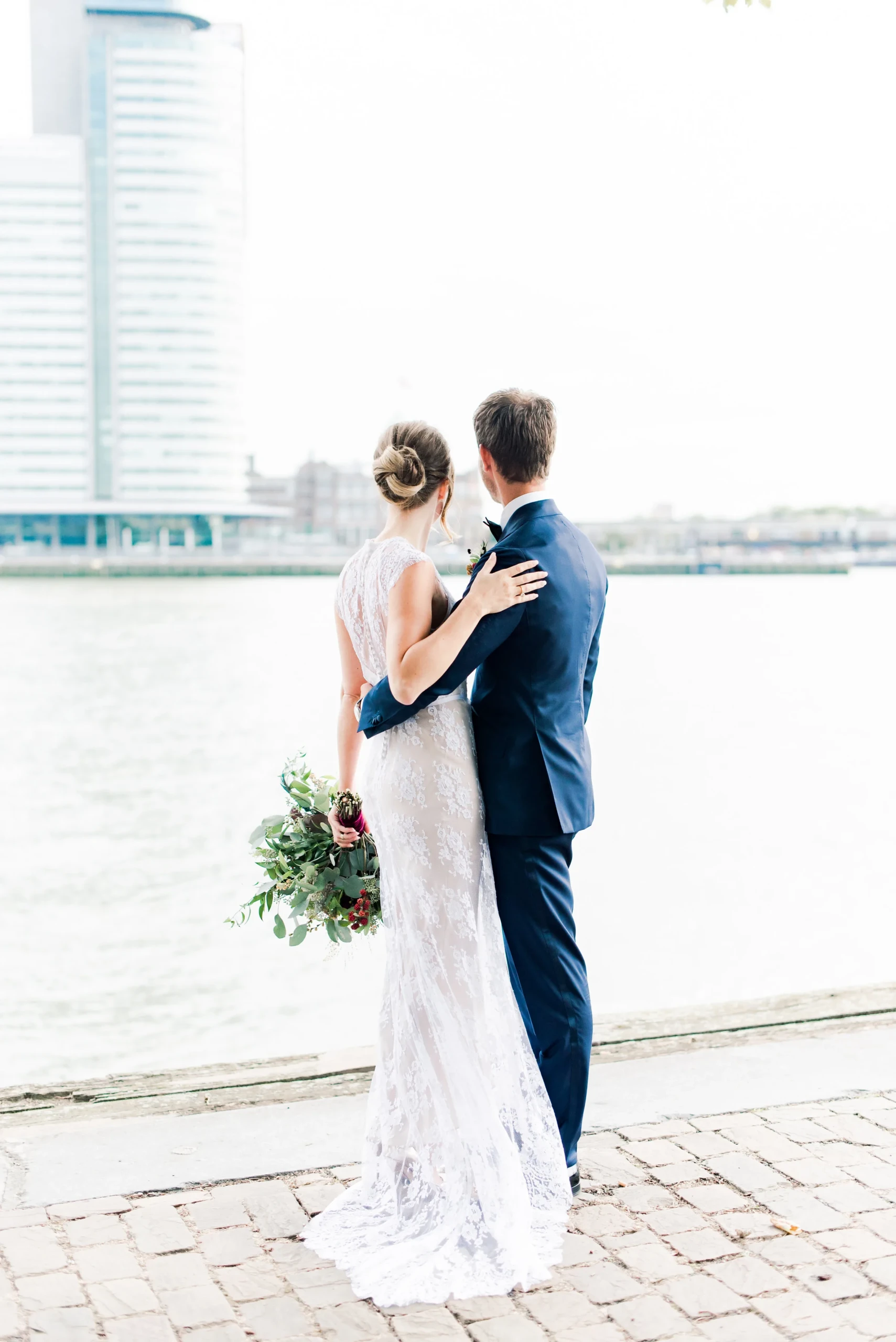 Bruid in witte trouwjurk met opgestoken haar staat met de bruidegom uit te kijken over de Maas naar Hotel New York in Rotterdam