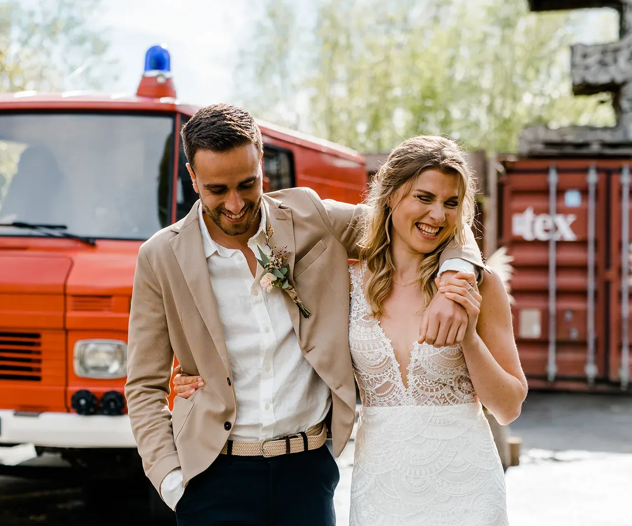 Lachend bruidspaar staan voor een oude Duitse brandweerwagen. Bruid draagt een witte trouwjurk en bruidegom een two-tone trouwpak inclusief corsage