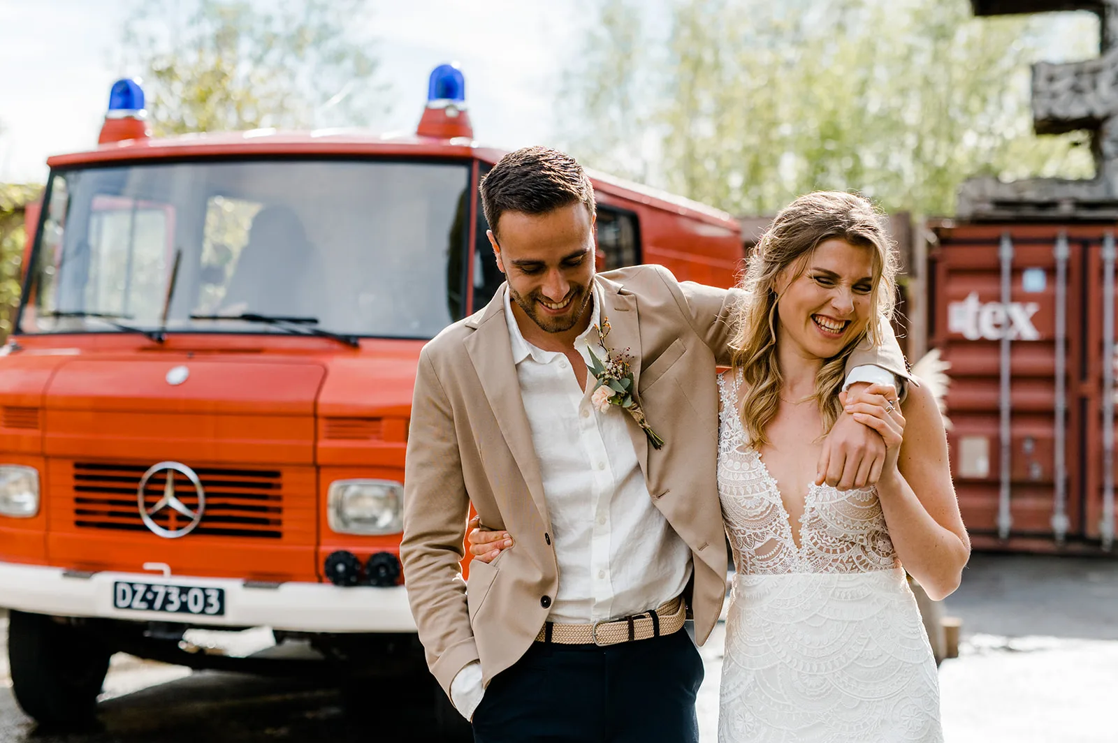 Lachend bruidspaar staan voor een rode brandweerwagen. Bruid draagt een kante trouwjurk en bruidegom een bruin jasje met een donkerblauwe broek
