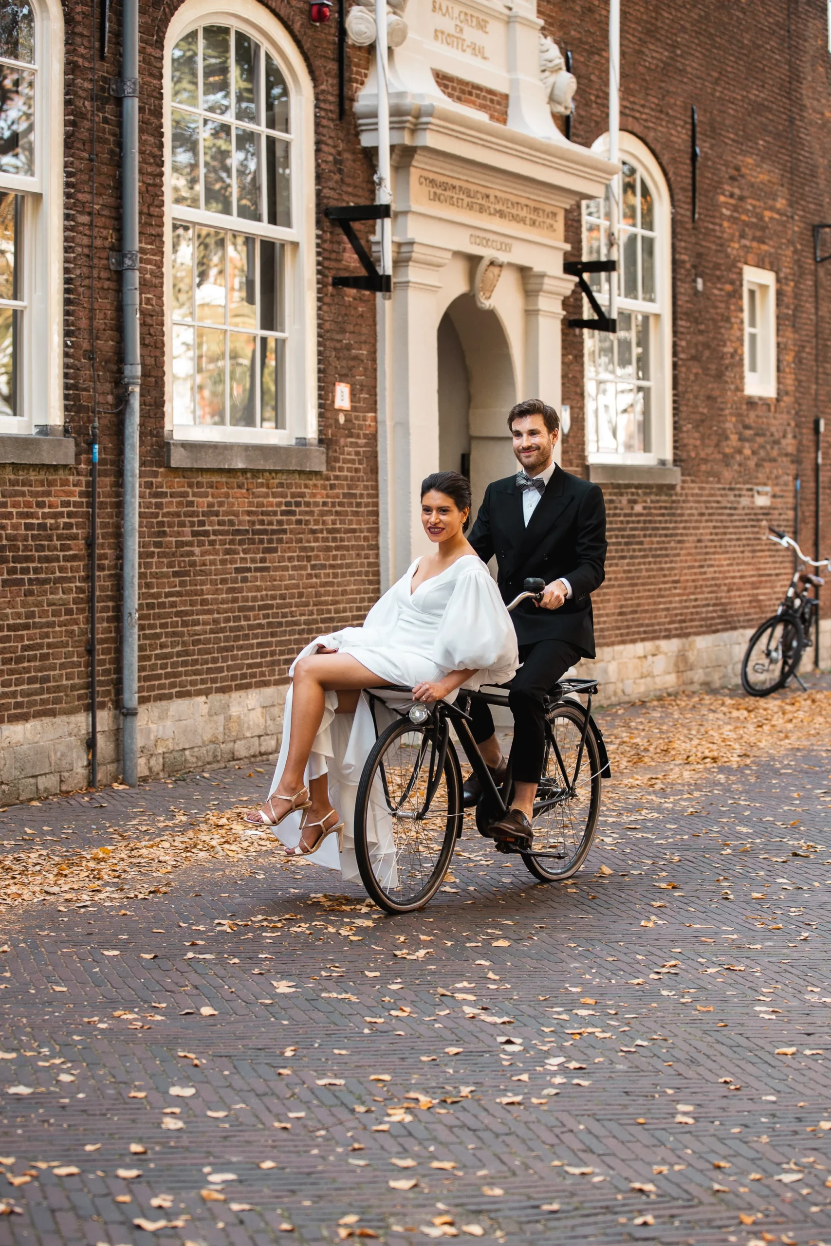 De bruid zit met haar witte bruidsjurk met hoge spilt voorop de fietsendrager en de bruidegom in een zwart pak met vlinderdas fietst
