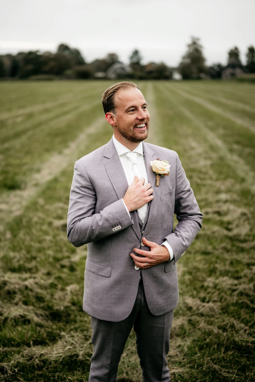Een bruidegom staat lachend in een open grasveld met rijbanen in de achtergrond. Hij draagt een lichtgrijs pak met een witte das en een crèmekleurige roos als corsage. Hij houdt met één hand zijn jasje vast ter hoogte van zijn borst en kijkt trots voor zich uit.