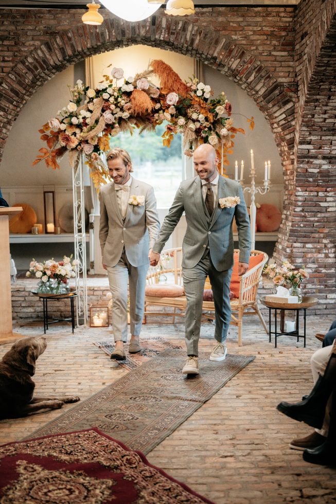 Twee bruidegoms lopen hand in hand door het gangpad van een sfeervolle trouwlocatie met bakstenen muren en een bloemenboog met herfsttinten. Ze dragen lichtgrijze pakken met corsages en stralen van geluk. Op de voorgrond ligt een bruine hond op een Perzisch tapijt, omringd door bloemen, kaarsen en gasten.