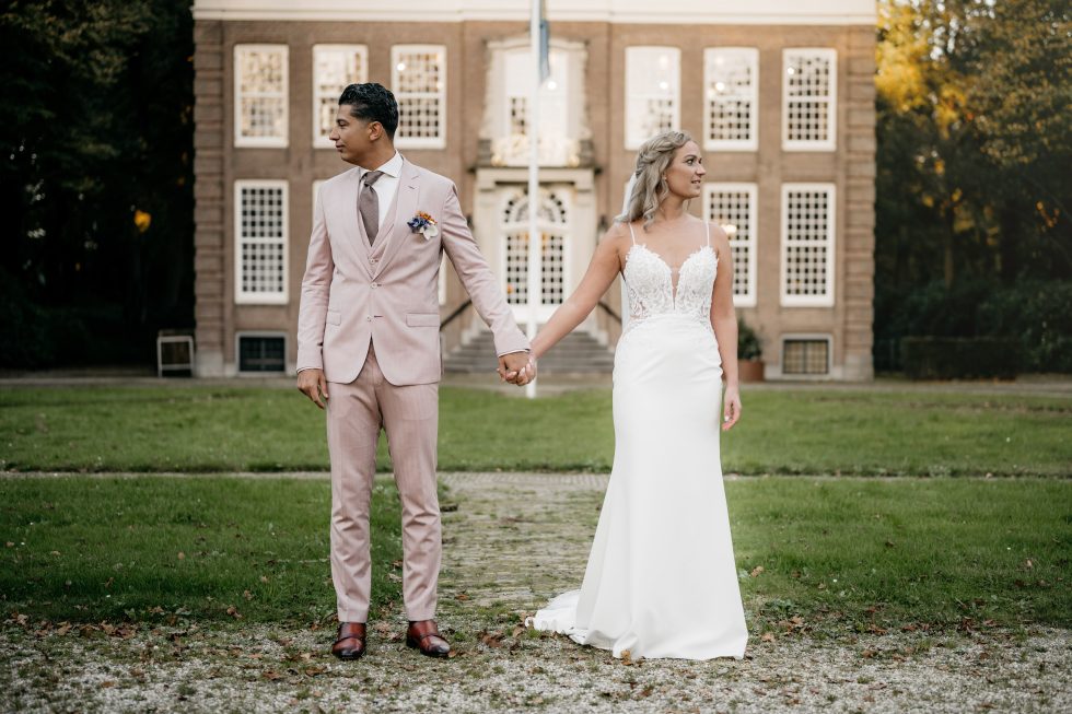 Een bruid en bruidegom staan hand in hand voor een statig landhuis met witte ramen. De bruid draagt een elegante witte jurk met kanten details en kijkt naar rechts, terwijl de bruidegom in een lichtroze pak met corsage naar links kijkt. Ze staan op een grindpad tussen het gras.
