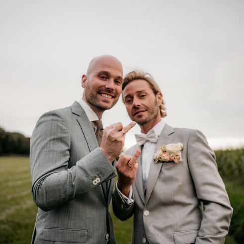 Twee mannen in lichtgrijze trouwpakken poseren vrolijk in een weiland, beiden dragen trouwringen. Ze maken speelse handgebaren naar de camera, de ene toont zijn middelvinger en de andere het rock-gebaar. Beiden lachen en stralen geluk uit, met bloemen op hun revers.