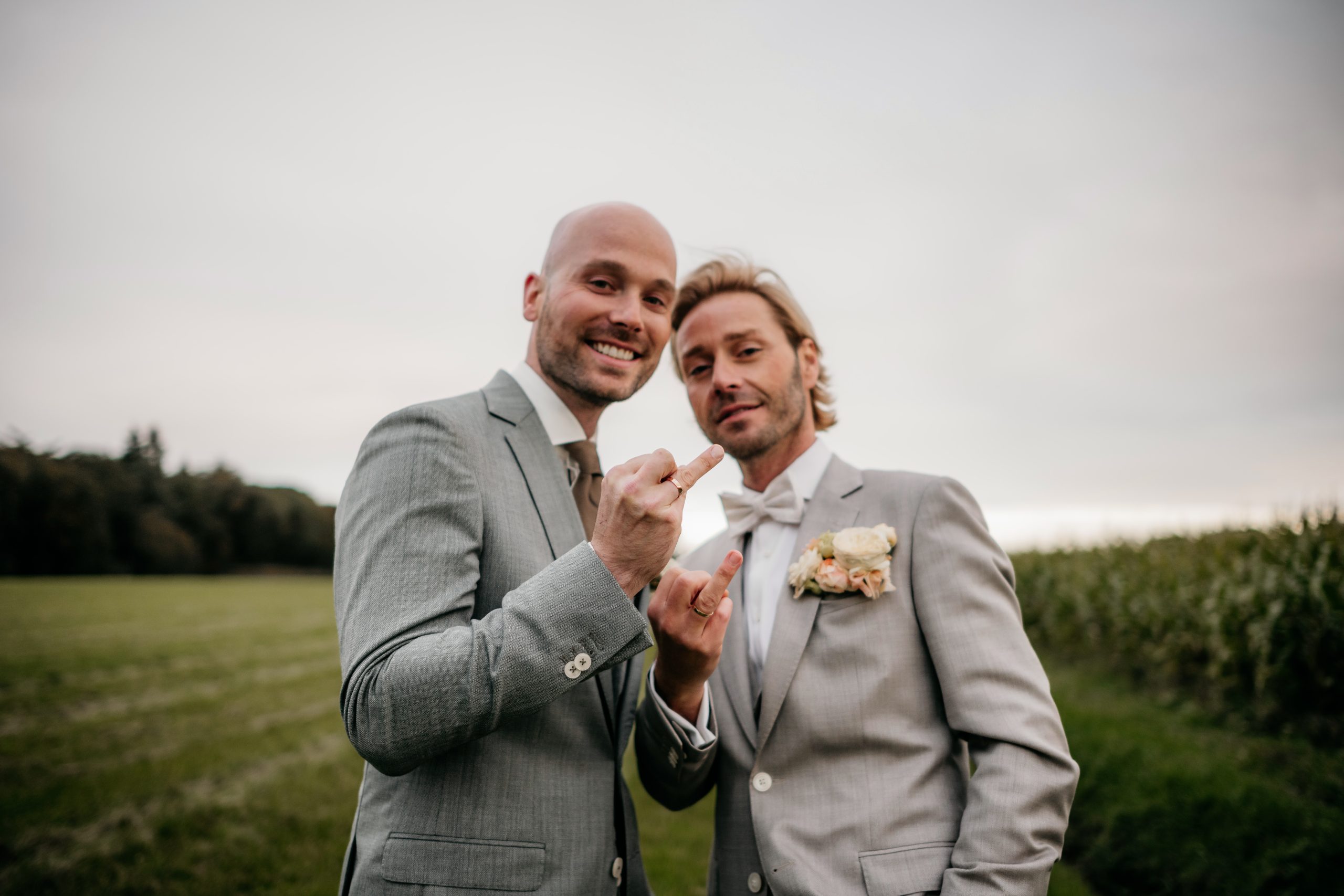 Twee mannen in lichtgrijze trouwpakken poseren vrolijk in een weiland, beiden dragen trouwringen. Ze maken speelse handgebaren naar de camera, de ene toont zijn middelvinger en de andere het rock-gebaar. Beiden lachen en stralen geluk uit, met bloemen op hun revers.