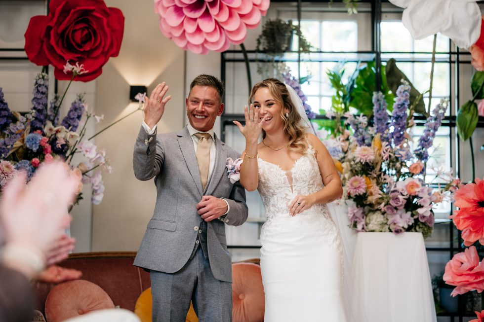 Een bruid en bruidegom tonen lachend hun trouwringen aan de gasten. Ze staan omringd door kleurrijke bloemen en grote papieren bloemen aan het plafond en de muren. De bruid draagt een kanten trouwjurk en de bruidegom een lichtgrijs pak met een gouden das.