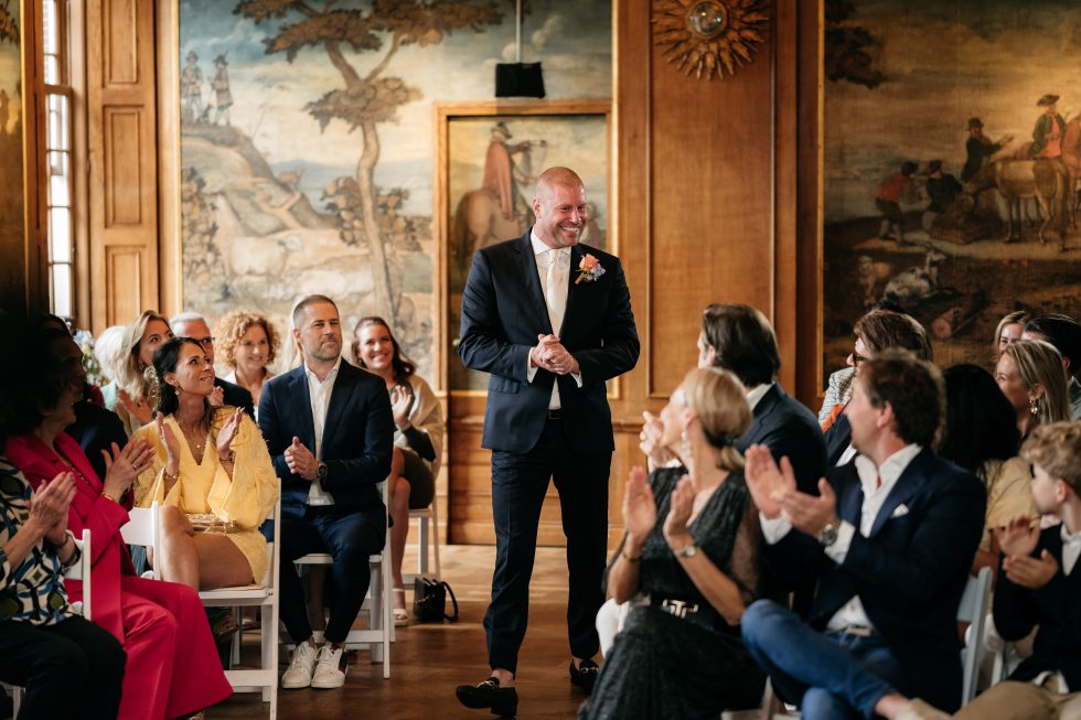 Een man in donkerblauw pak met corsage en witte das loopt lachend door het middenpad van een elegante trouwzaal, omringd door applaudisserende gasten die op witte stoelen zitten. De zaal is rijkelijk versierd met klassieke muurschilderingen en houten panelen.