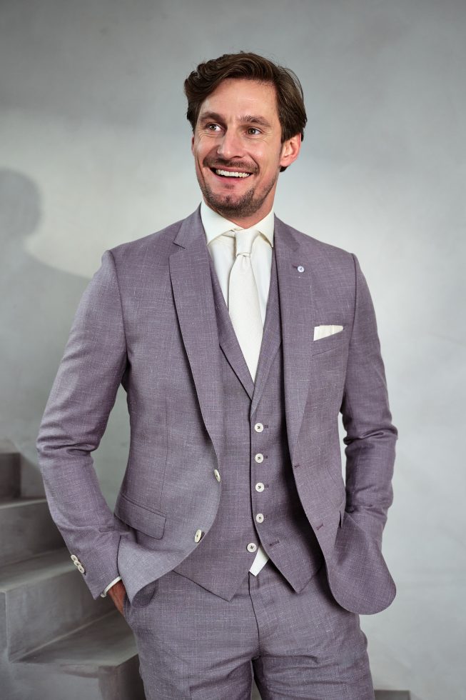 Man in lichtpaars driedelig pak en witte das poseert glimlachend met handen in de zakken tegen een lichtgrijze achtergrond.