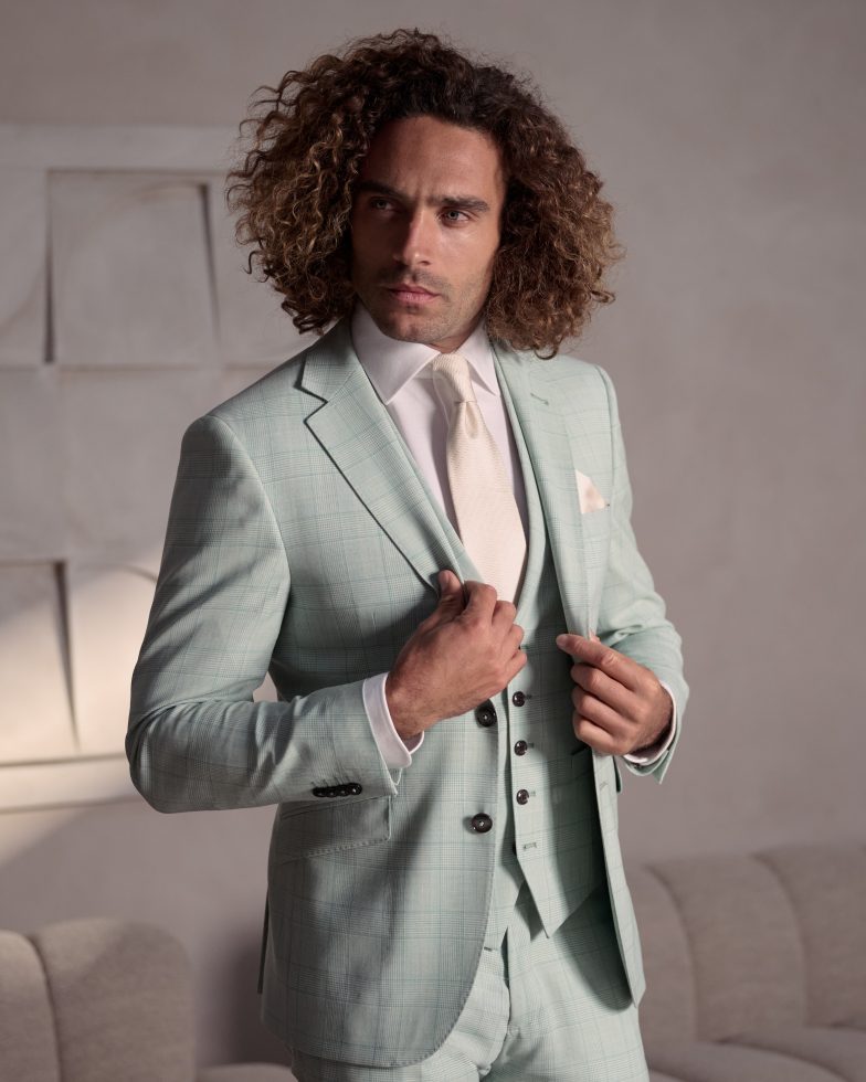 Dezelfde man draagt een mintgroen geruit pak met wit overhemd en crèmekleurige das, en kijkt serieus terwijl hij zijn jasje rechttrekt.