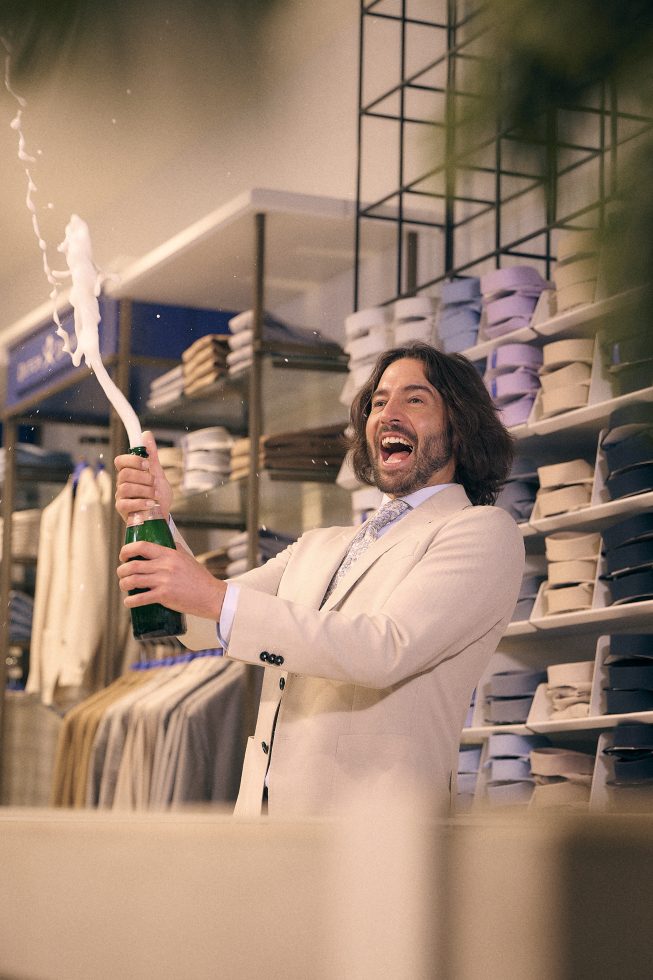 Man in crème pak en bloemetjesdas opent een champagnefles met een grote schuimexplosie in een stijlvolle winkel vol overhemden en pakken.