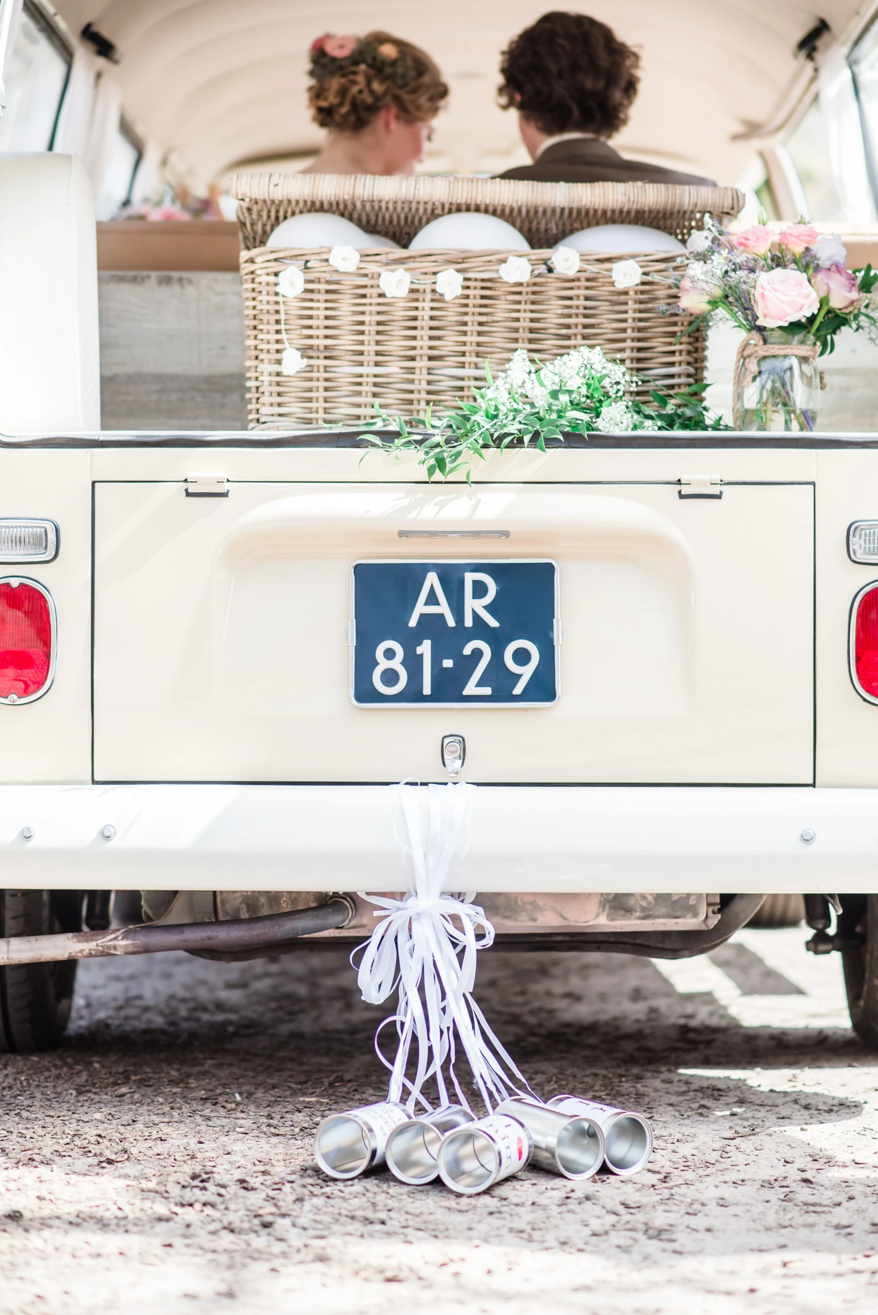 de achterkant van een Volkswagen bus met daarin het bruidspaar. Aan de bumper hangen 5 blikjes. In de kofferbak staat een rieten mand met witte ballonnen en een boeket in een glazen potje