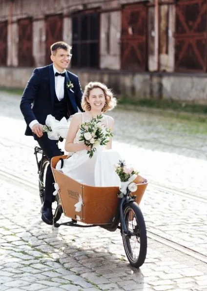 Bruid zit in een bakfiets met haar bruidsboeket. Bruidegom in donker driedeling trouwpak fietst.