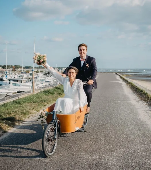 Bruid zwaait vanuit de bakfiets en bruidegom fietst van de dijk af. Naast de dijk ligt een haven met boten en aan de andere kant ligt het strand.