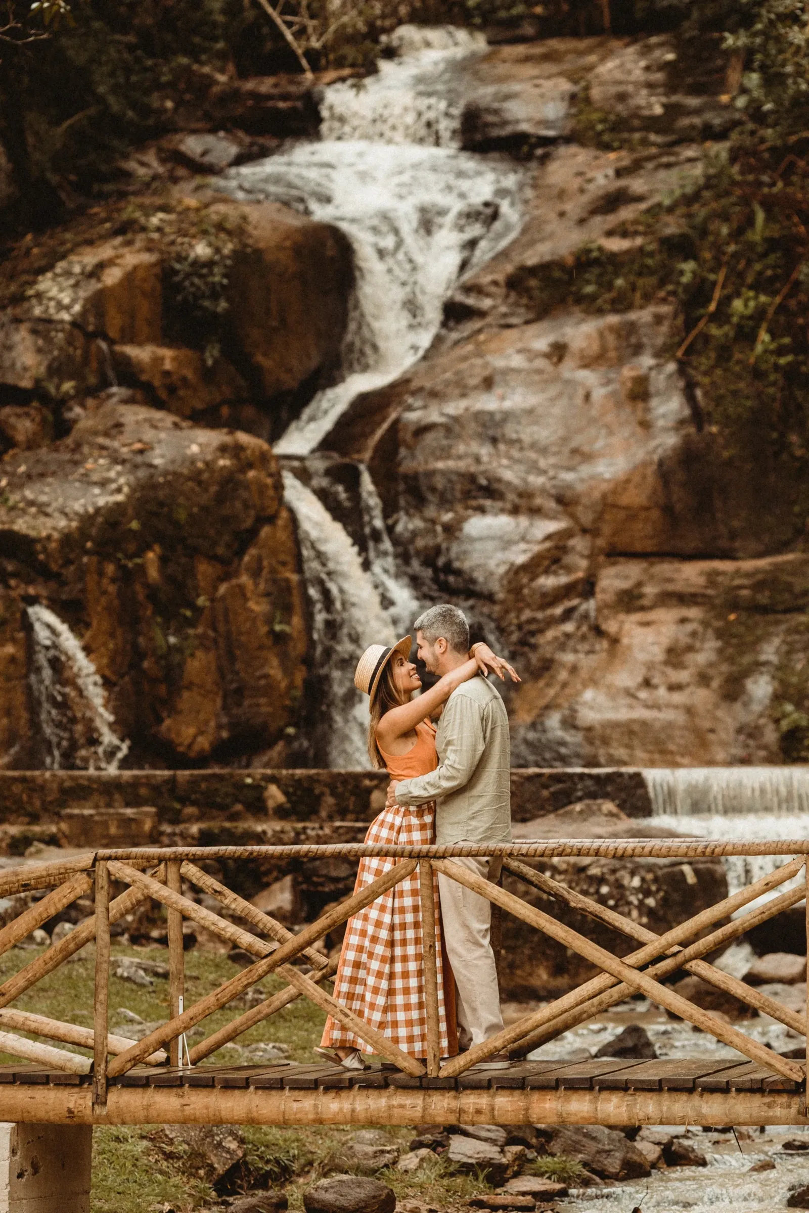 Verliefd stel staat op een houten brug voor een waterval en omhelzen elkaar. Vrouw draagt een rieten hoed, oranje top en lange geruite jruk. Man draagt een zandkleurige outfit