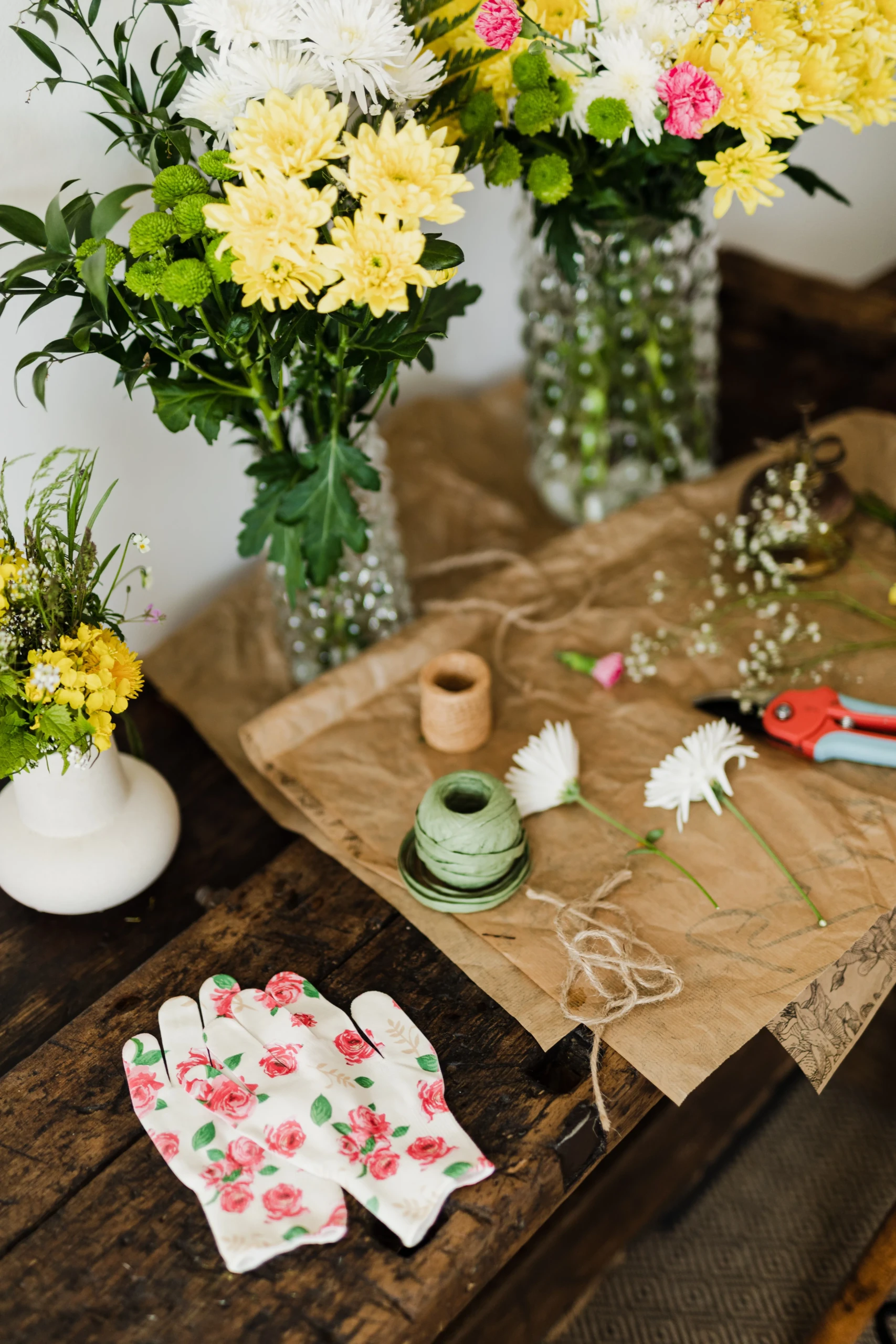 Op een houten werkbank liggen handschoenen met bloemmotief, een rose-blauwe bloemenschaar, en gele, witte een roze bloemen in drie vazen.