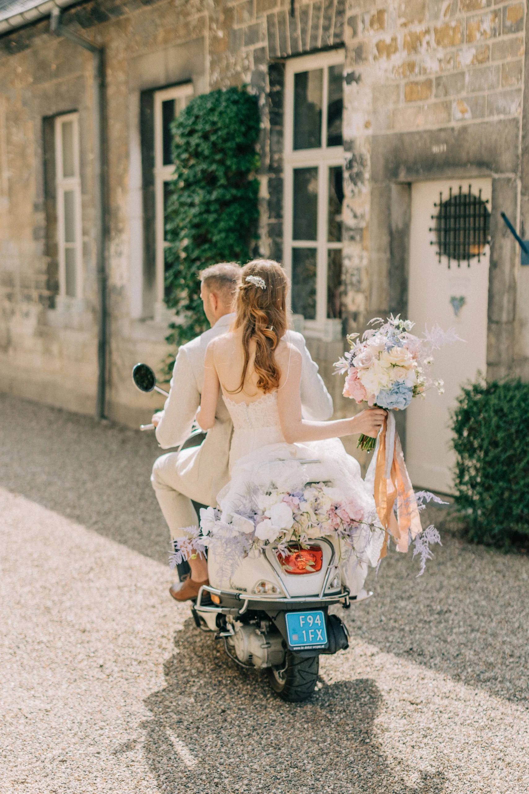 Bruidspaar rijdt weg over een grindpad op een met bloemen versierde scooter. Achterop zit de bruid met een groot pastelkleurig bruidsboeket