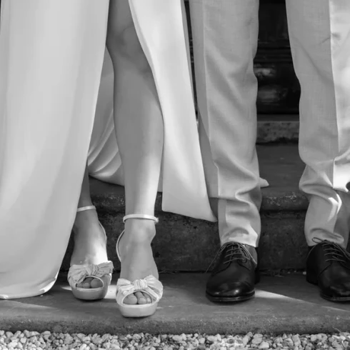 de benen van de bruid met open trouwschoenen en een lange split aan de voorkant van de trouwjurk. En de twee benen van de bruidegom met gepoetste herenmode schoenen.