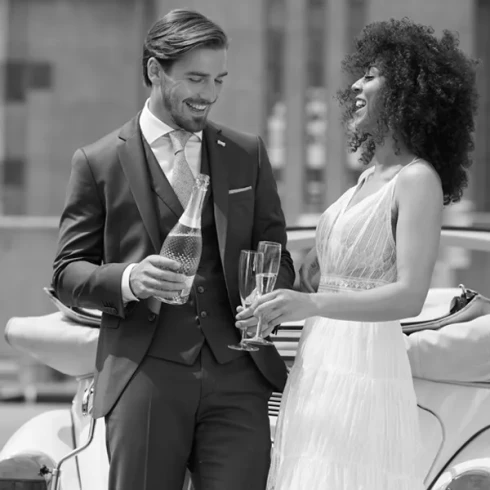 De bruid houdt twee champagne glazen vast en de bruidegom houdt de champagnefles vast. Bruidspaar staat lachend voor een Volkswagen Kever Cabrio.