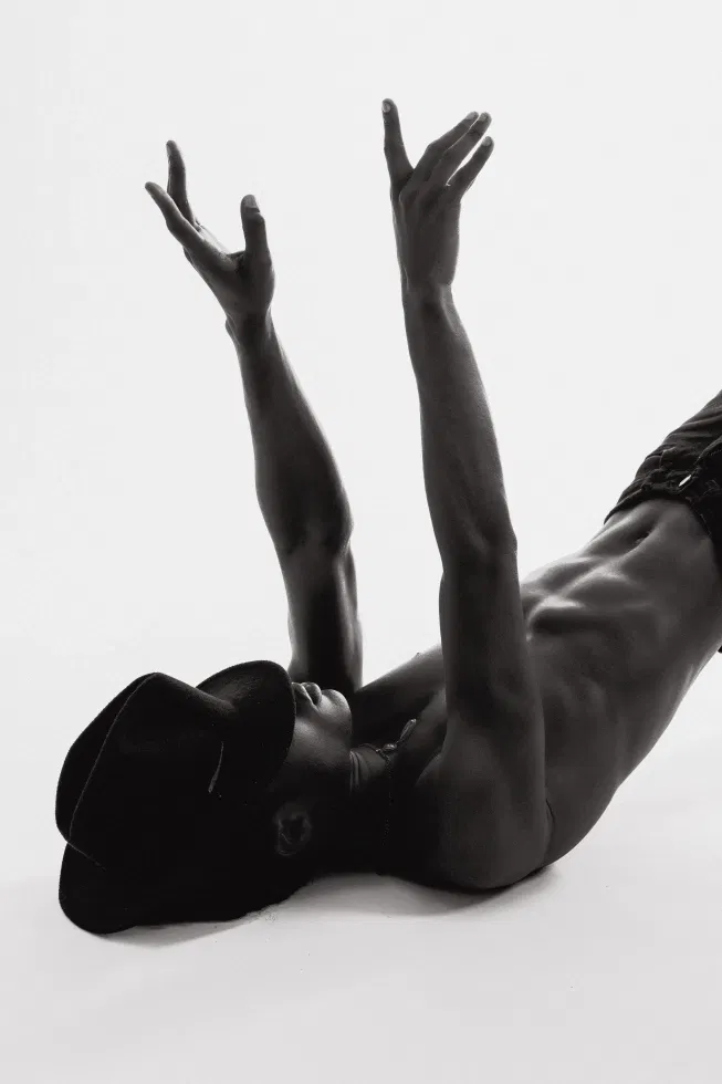 Zwartwit foto. Man met ontbloot bovenlichaam en jeans aan. Ligt met zijn bovenlichaam op de grond en heeft een hoed over zijn gezicht.